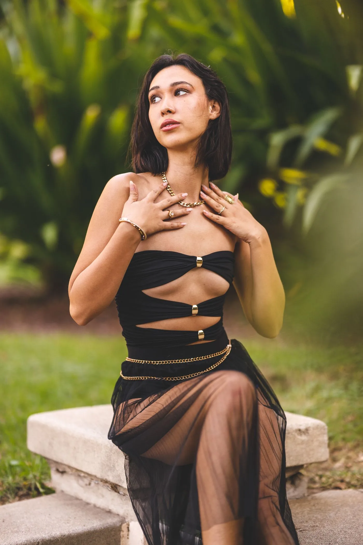 A woman in a dark cutout top and a long brown skirt sitting beside greenery.