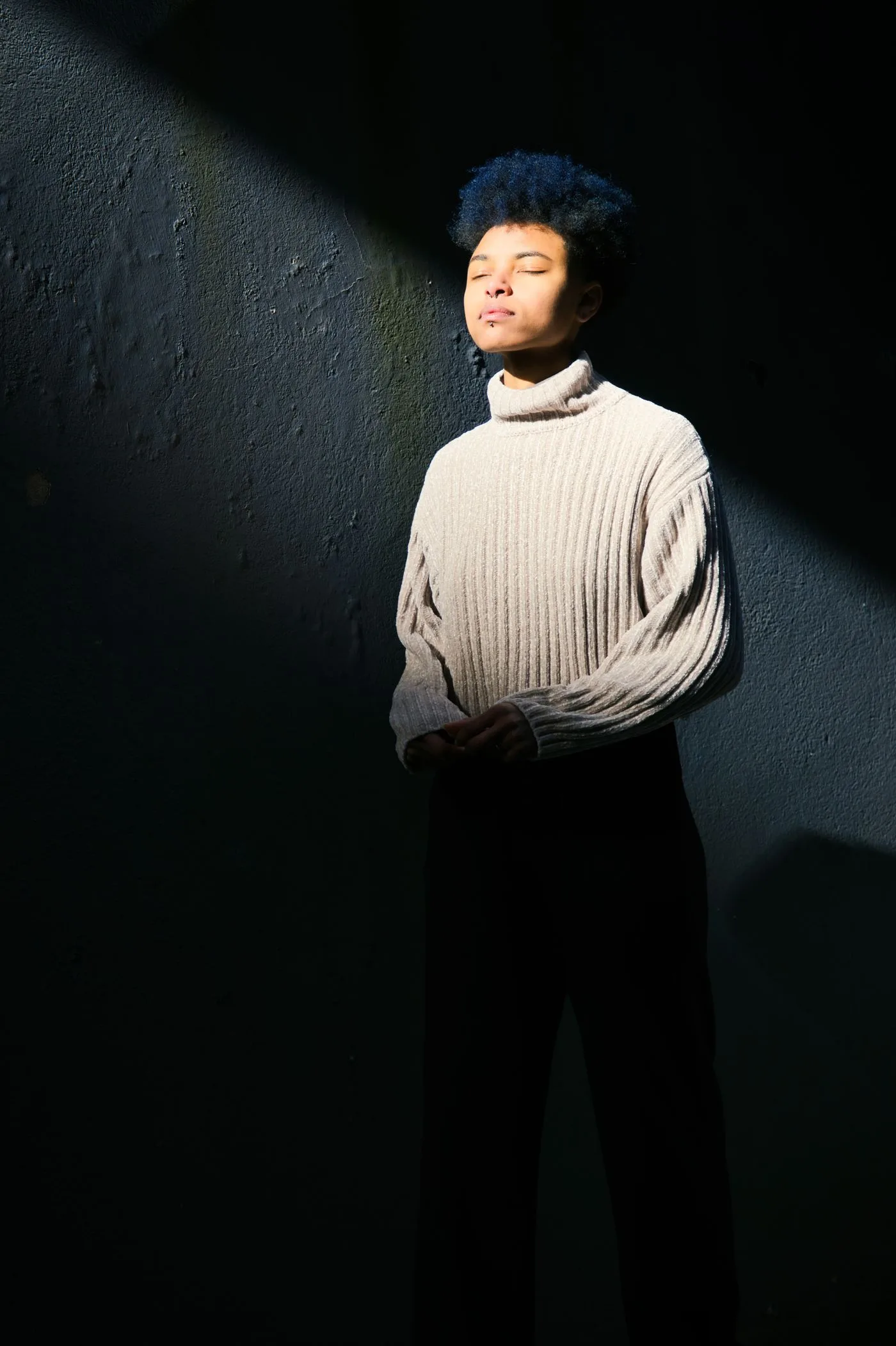 A person in a gray turtleneck sweater standing in sunlight near a wall.