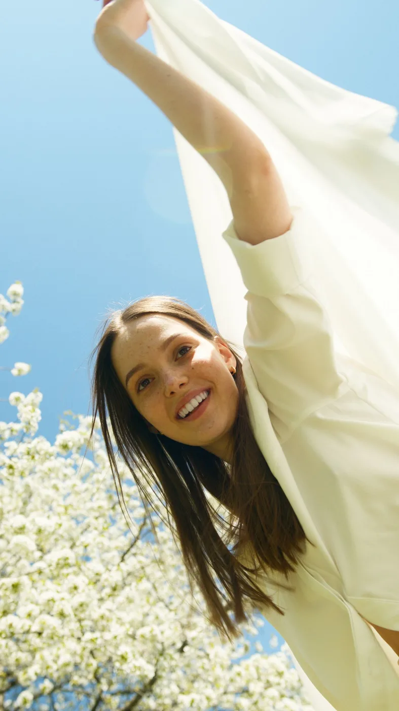 A woman raising the sleeve of a light flowing garment outdoors beneath a blue sky.