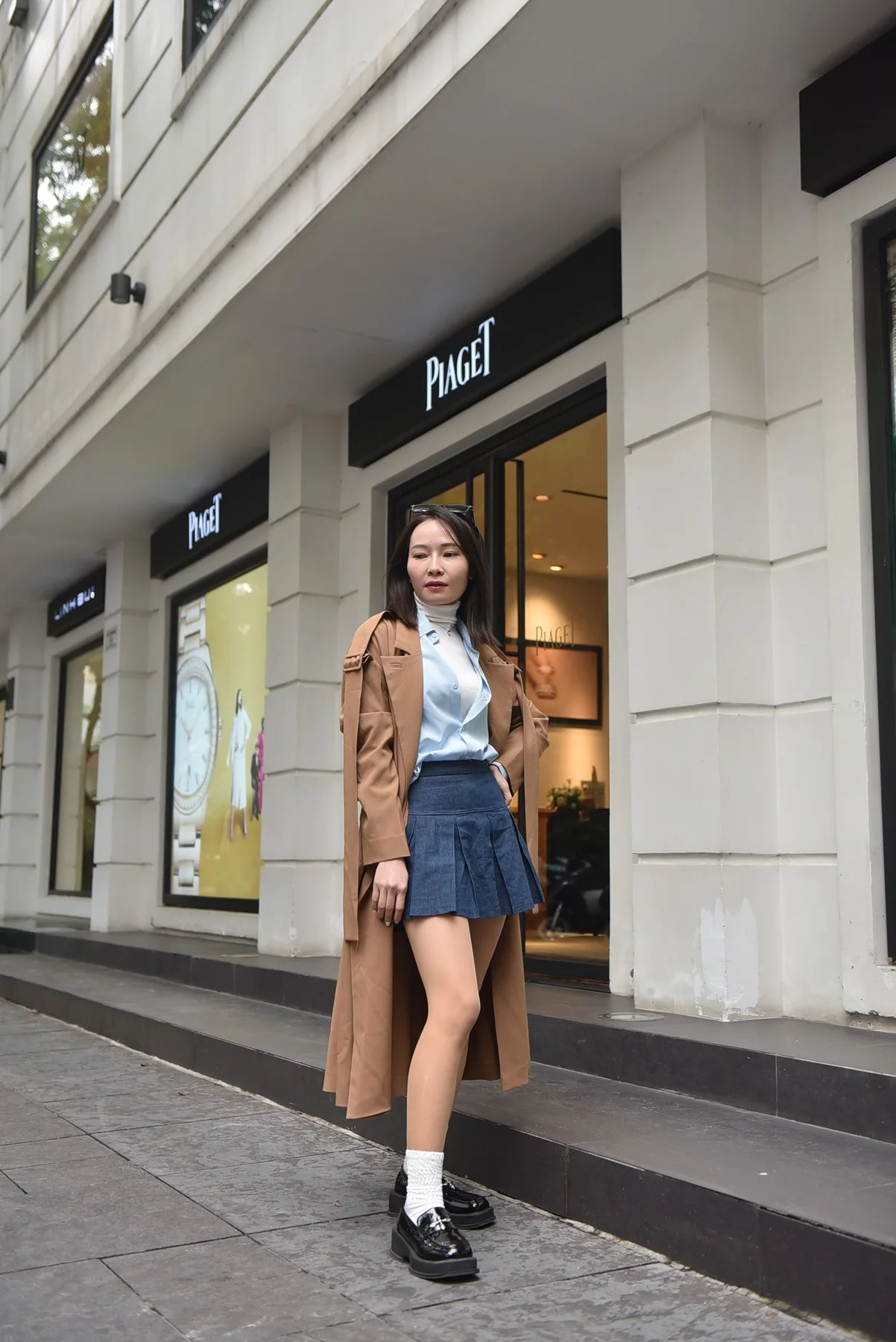 A woman wearing a brown long coat with a blue pleated skirt and dark shoes