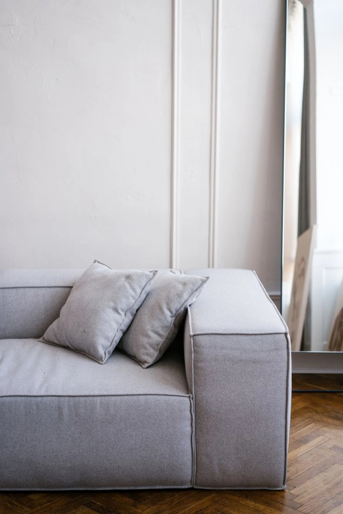 A grey sofa with two cushions beside a tall mirror and pale wall