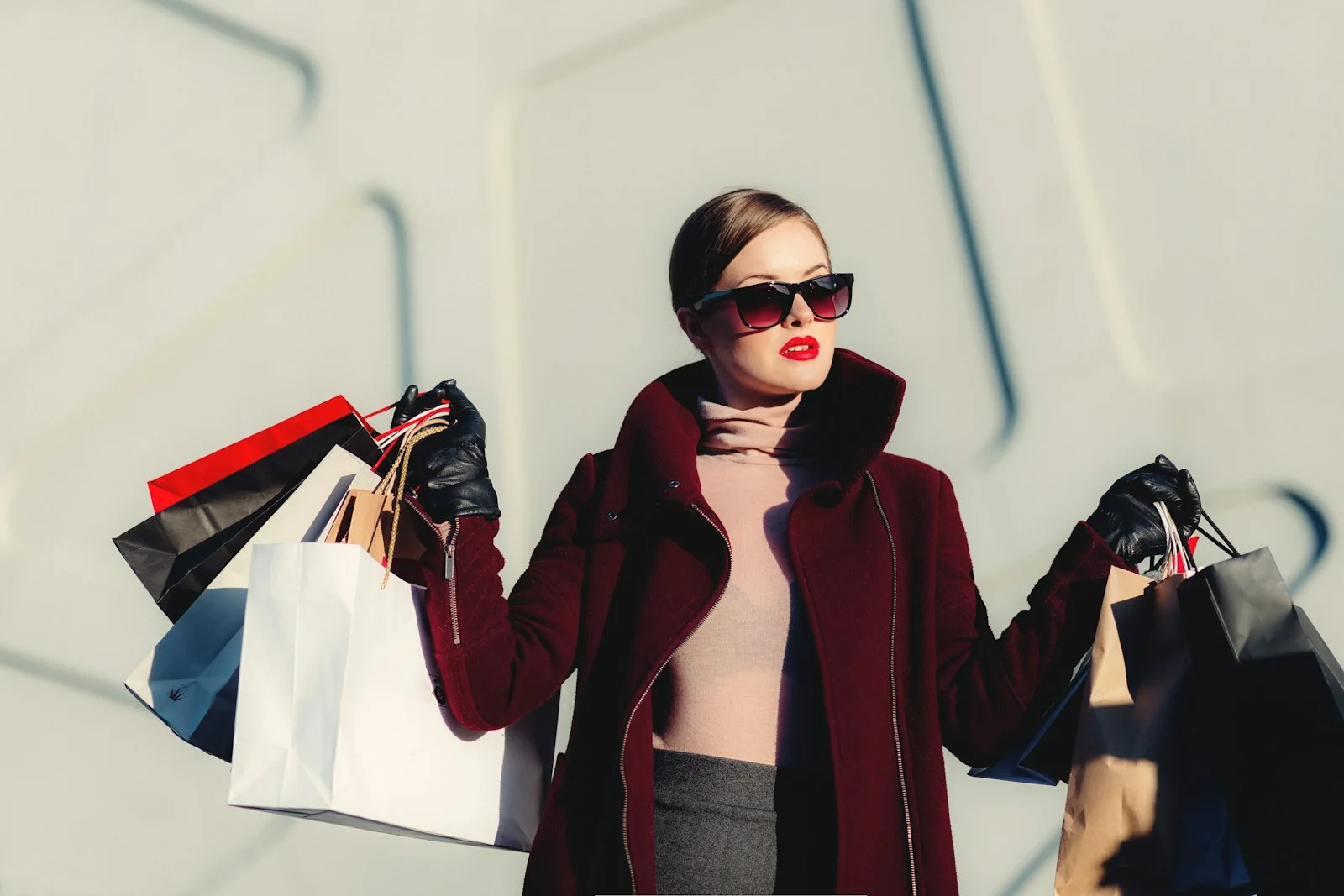 A woman wearing a burgundy coat and sunglasses holds shopping bags