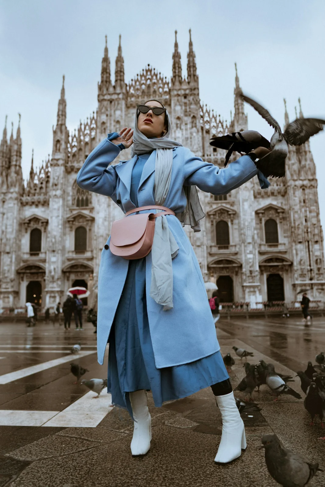 A woman wears a blue belted trench-style coat and carries a pink shoulder bag