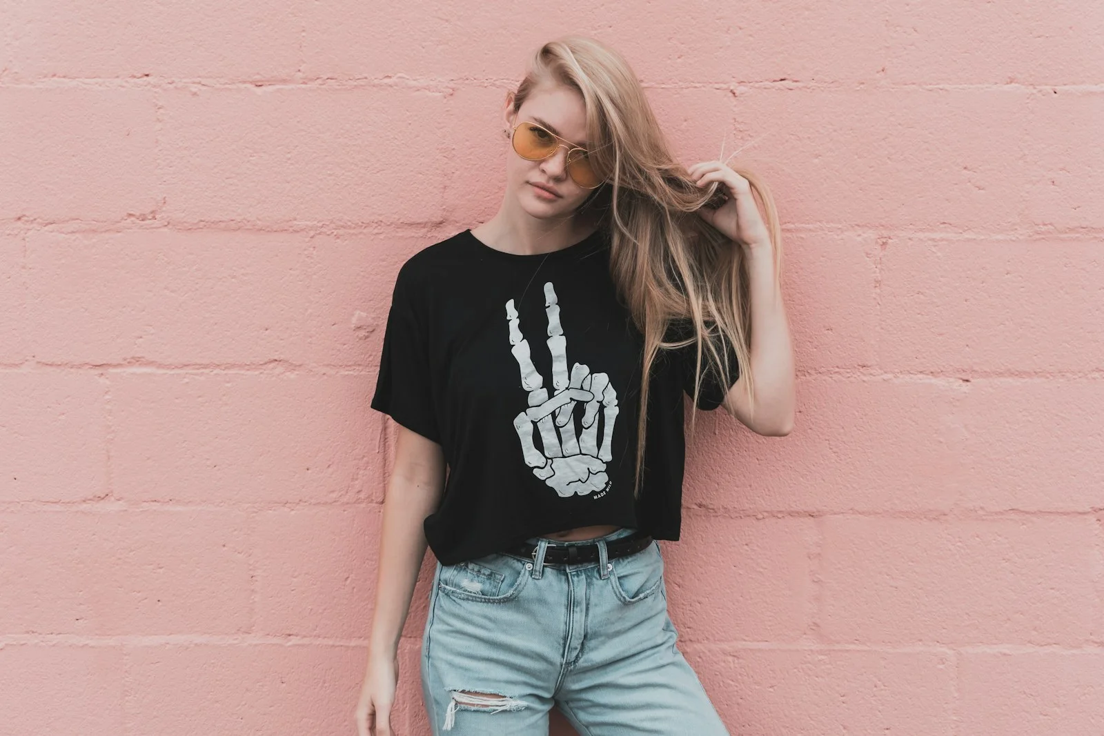 A woman wears a black graphic T-shirt and pale blue jeans against a pink wall